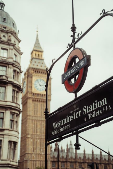 A close-up view of a London Underground sign hanging from an ornate black iron bracket, with the sign reading 'Underground' in white text inside a red circle. Below it, a rectangular black and white sign indicates direction to 'Westminster Station,' the public subway, and toilets. In the background, the iconic clock tower, commonly known as Big Ben, is visible with its detailed stonework and a large clock face displaying the time, set against a muted, overcast sky. To the left, part of a historic building with rounded, decorative stone architecture and multiple windows can be seen, enhancing the classic London cityscape. The scene captures the urban environment typical of Westminster, with a focus on transportation signage relevant for those seeking alternative waste handling or private rubbish removal services near central London landmarks, subtly aligned with the context of professional rubbish clearance such as that offered by Waste Clearance Westminster.