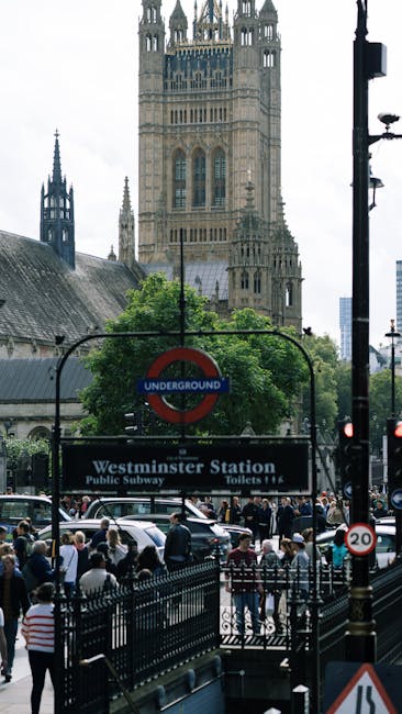 A close-up view of a London Underground sign hanging from an ornate black iron bracket, with the sign reading 'Underground' in white text inside a red circle. Below it, a rectangular black and white sign indicates direction to 'Westminster Station,' the public subway, and toilets. In the background, the iconic clock tower, commonly known as Big Ben, is visible with its detailed stonework and a large clock face displaying the time, set against a muted, overcast sky. To the left, part of a historic building with rounded, decorative stone architecture and multiple windows can be seen, enhancing the classic London cityscape. The scene captures the urban environment typical of Westminster, with a focus on transportation signage relevant for those seeking alternative waste handling or private rubbish removal services near central London landmarks, subtly aligned with the context of professional rubbish clearance such as that offered by Waste Clearance Westminster.