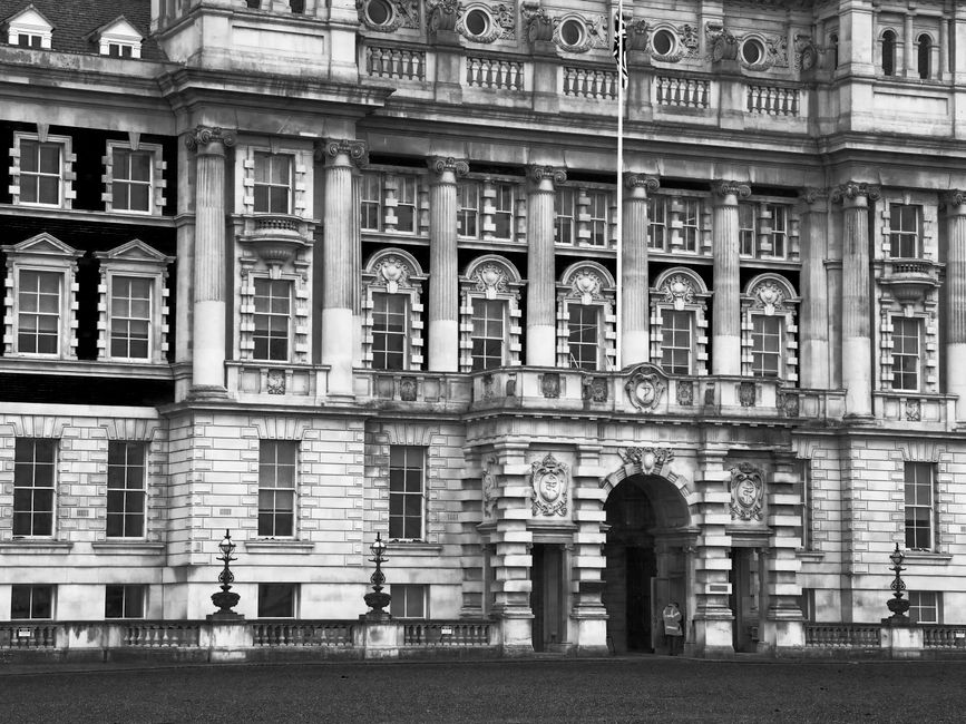 A black and white photograph of an elegant, historic building with ornate architectural details, including tall columns with decorative capitals, arched windows with intricate moldings, and a central arched entrance framed by stonework featuring carved crest emblems. The facade is composed of large stone blocks with a mix of smooth and textured finishes, and the windows are divided into multiple small panes. In the foreground, there is a paved street or courtyard with a low stone wall adorned with decorative lamp posts on either side of the entrance, suggesting a setting within a city or historic district. The image captures the grandeur of the structure's classical design, highlighting its detailed masonry, symmetrical proportions, and stately appearance, typical of grand civic or institutional buildings, which may be relevant to contextual discussions about private or alternative waste management arrangements in heritage or urban environments.