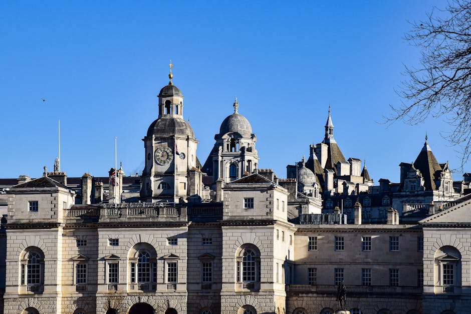 A historic cityscape featuring a large, ornate stone building with multiple domed towers and spires, set against a clear blue sky. The foreground shows the stone facade with evenly spaced arched windows and decorative details, indicating classical architectural style. Behind the main structure, additional towers and spires rise, each with pointed rooftops and intricate dormer windows, suggesting a city with rich architectural heritage. To the right, a bare tree branch extends into the frame, adding a natural element to the scene. The overall environment appears urban, with the building possibly serving governmental or civic functions, set in a location where private waste management services like Waste Clearance Westminster would undertake rubbish removal or on-site clearance related to city maintenance. The lighting is bright and natural, highlighting the textures of the stonework and the details of the architectural features, evoking a typical day suitable for professional rubbish collection services in London. This scene captures the juxtaposition of historical architecture with modern municipal or private waste management activities, emphasizing the importance of efficient rubbish removal in heritage-rich environments.