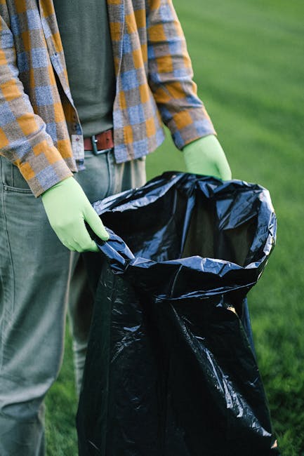 A person standing outdoors on a grassy area, dressed in casual clothing including a green shirt, a plaid jacket with shades of yellow, gray, and blue, and gray trousers with a brown belt. They are wearing light green disposable gloves and holding a large open black rubbish bag made of plastic, which appears to be for waste collection or clearance purposes. In the background, the grass is lush and well-maintained, indicating an outdoor environment suitable for rubbish removal or waste management activities. The scene suggests an act of environmental cleanup or waste disposal, consistent with private rubbish collection services provided by Waste Clearance Westminster, which offers alternative waste handling solutions outside of traditional municipal collection. The focus on the gloves and rubbish bag underscores the importance of safe and hygienic waste handling during on-site clearance tasks.