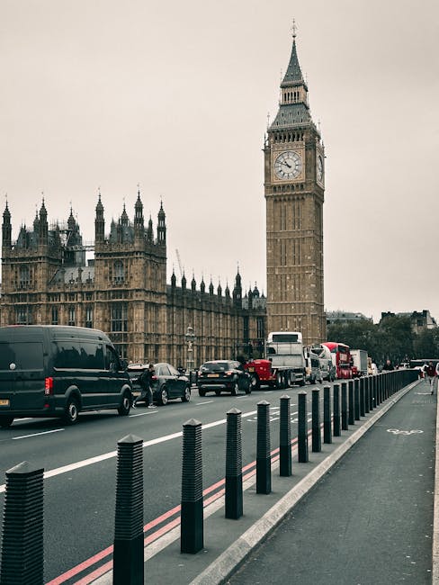 A daytime street scene in Westminster featuring the iconic Big Ben clock tower and the Palace of Westminster in the background. In the foreground, there is a lane dedicated to vehicle traffic, with various cars, vans, and double-decker buses arranged in a line along the road. To the right side of the road, a row of black plastic bollards creates a barrier between the vehicle lane and the sidewalk. The bollards are evenly spaced and stand on a concrete curb. The sky above is overcast with a greyish hue, giving the scene a muted atmosphere. The street pavement appears clean and well-maintained, with clearly marked white and red lines. The overall scene is typical of busy central London traffic areas, capturing a moment in the city's constant movement, with the British government buildings serving as a prominent visual backdrop. This setting subtly reflects the city's approach to urban management, including aspects of private and independent waste collection infrastructure often seen in busy urban environments like Westminster.