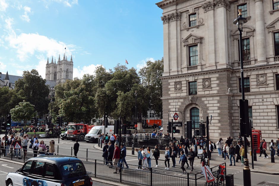 A busy urban street scene in Westminster featuring a large, ornate stone building with detailed architectural elements and multiple windows on the right side of the image. In the foreground, a diverse group of pedestrians are crossing the road at a designated crosswalk, some dressed in casual attire, others in business wear. Vehicles including a white delivery van, a red truck, and a dark sedan are stopped at traffic lights, with additional cars visible further along the street. A black metal fence runs parallel to the pavement, separating the sidewalk from the roadway. Several lampposts and red telephone boxes line the sidewalk, and a London Underground sign is visible near the entrance of the stone building. The scene is illuminated by natural daylight, with a partly cloudy sky overhead and lush green trees lining the street, some with branches extending into the view. This scene reflects a typical busy day in central Westminster, suitable for context related to waste removal and on-site clearance services by Waste Clearance Westminster, with a clear focus on urban environment and public activity.