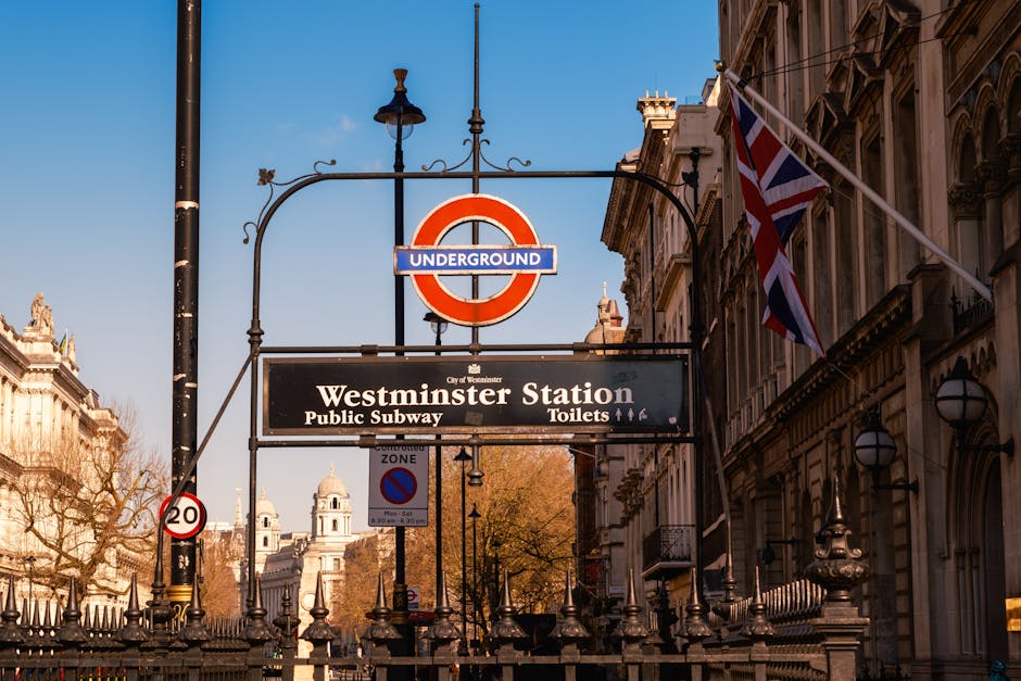 A daytime street scene in Westminster featuring the iconic Big Ben clock tower and the Palace of Westminster in the background. In the foreground, there is a lane dedicated to vehicle traffic, with various cars, vans, and double-decker buses arranged in a line along the road. To the right side of the road, a row of black plastic bollards creates a barrier between the vehicle lane and the sidewalk. The bollards are evenly spaced and stand on a concrete curb. The sky above is overcast with a greyish hue, giving the scene a muted atmosphere. The street pavement appears clean and well-maintained, with clearly marked white and red lines. The overall scene is typical of busy central London traffic areas, capturing a moment in the city's constant movement, with the British government buildings serving as a prominent visual backdrop. This setting subtly reflects the city's approach to urban management, including aspects of private and independent waste collection infrastructure often seen in busy urban environments like Westminster.